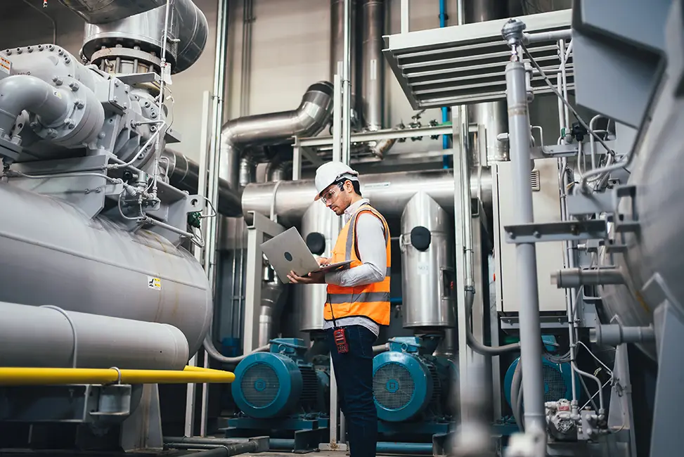 Engineer in an safety vest and hard hat looking at his laptop while inspecting/standing by a system of industrial pipes and machinery in a mechanical room.